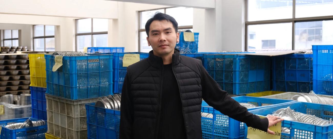 Retrato de Ying Changbin en su planta de producción de utensilios de cocina, rodeado de materiales y componentes.