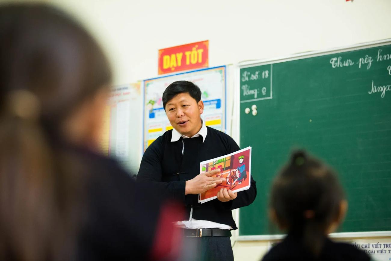 An image of a teacher pointing out something in a textbook in front of a class of students.
