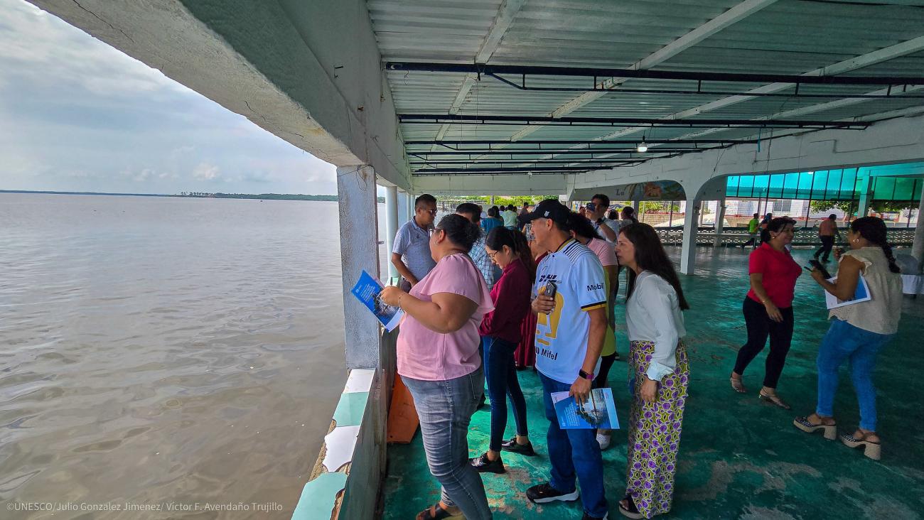 Un grupo de personas, de pie en una pasarela cubierta, observa el mar mientras sostiene cuadernos en las manos.