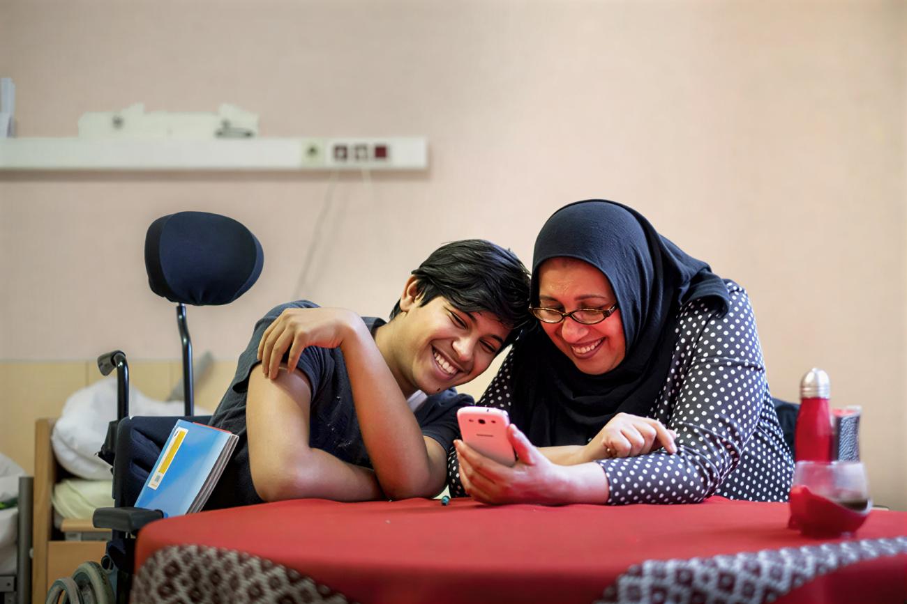 A boy and his mother laugh while watching something on her phone.