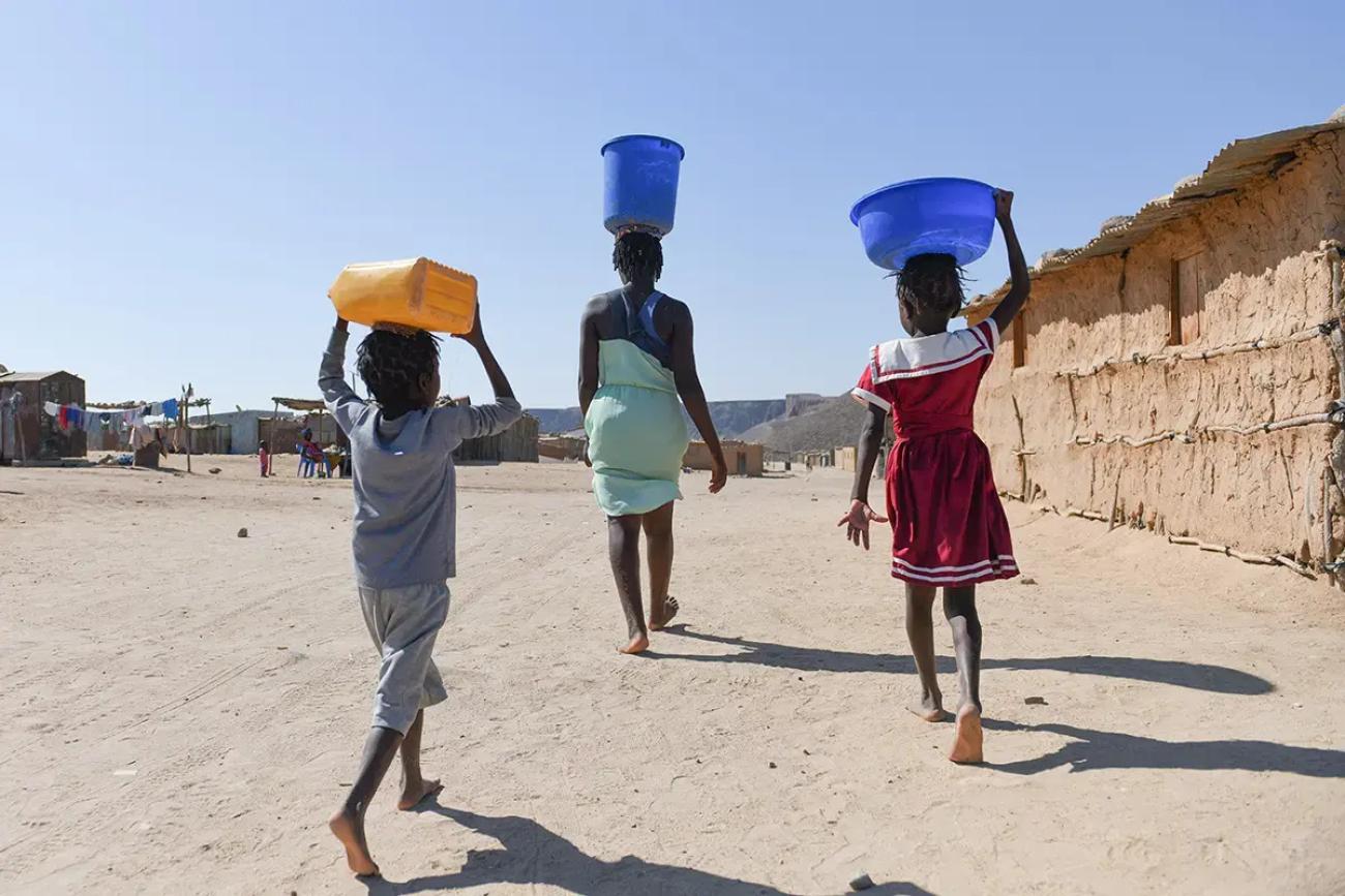 A woman in Angola walks with a bucket of water on her head, followed by her son and daughter, also carrying buckets.