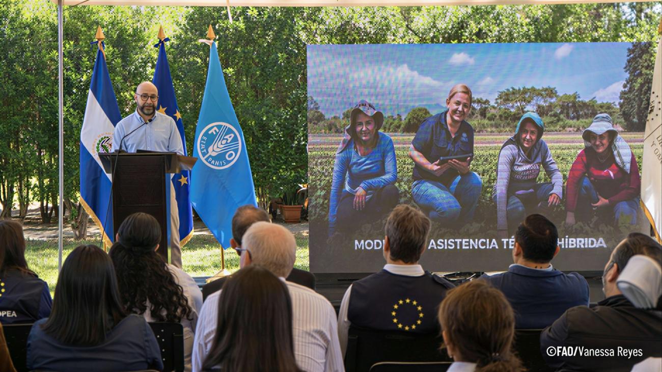 Un hombre da un discurso ante un público, junto a banderas y una pantalla con una imagen sobre trabajo agrícola.