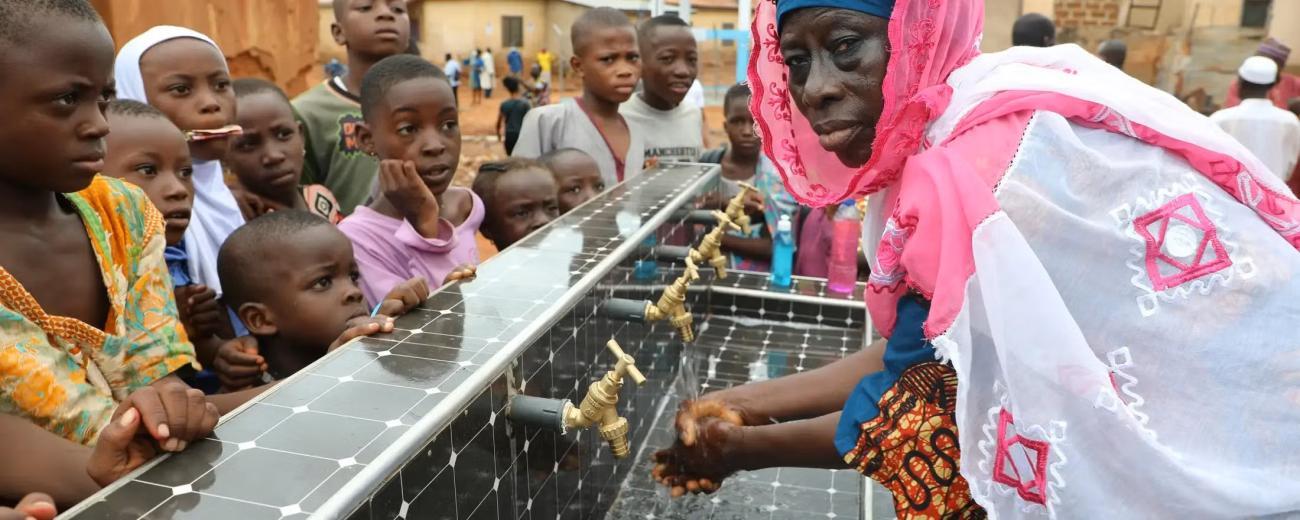 A woman in a pink dress washes her hands in a solar powered tap while children look on.