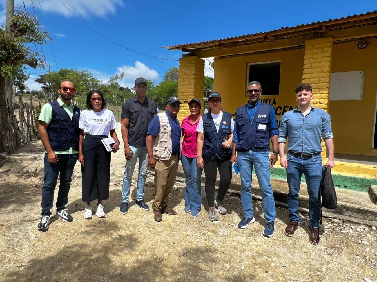 Representantes de organizaciones internacionales y autoridades locales visitan una comunidad rural frente a un edificio comunitario.