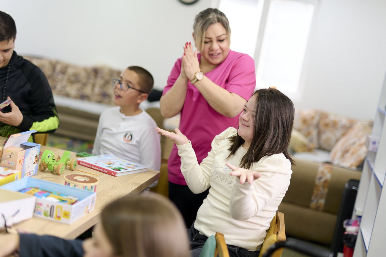 Group of people seated around a table engaging with puzzles and activities while one participant gestures and another claps nearby.