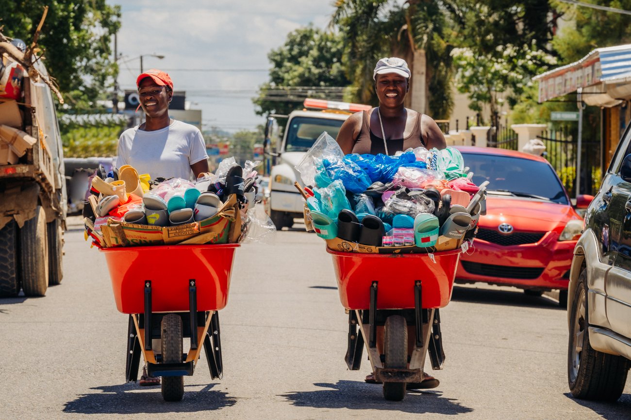 Two street vendors push red wheelbarrows filled with assorted footwear and goods along a busy road lined with vehicles and buildings.