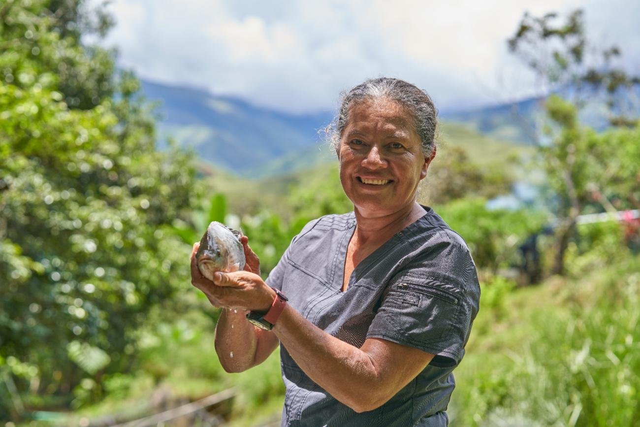 Una mujer sonriente sostiene un fruto entre las manos en un entorno rural montañoso y rodeado de vegetación.