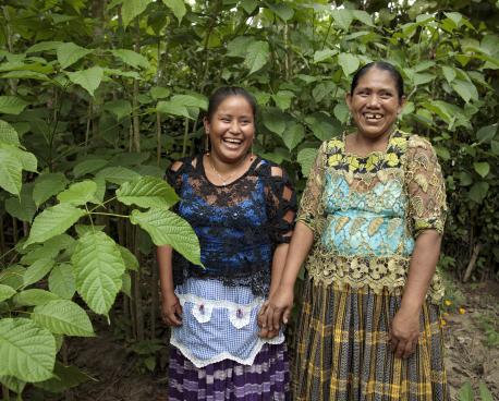 Deux femmes issues d’une communauté autochtone d’Amérique latine se tiennent debout, côte à côte, face caméra, au milieu d’une forêt, et arborent un large sourire. 