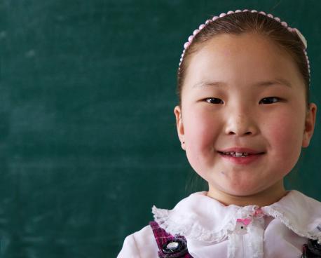 Muestre a una pequeña niña mongol sonriendo a la cámara mientras se para frente a una pizarra en blanco.
