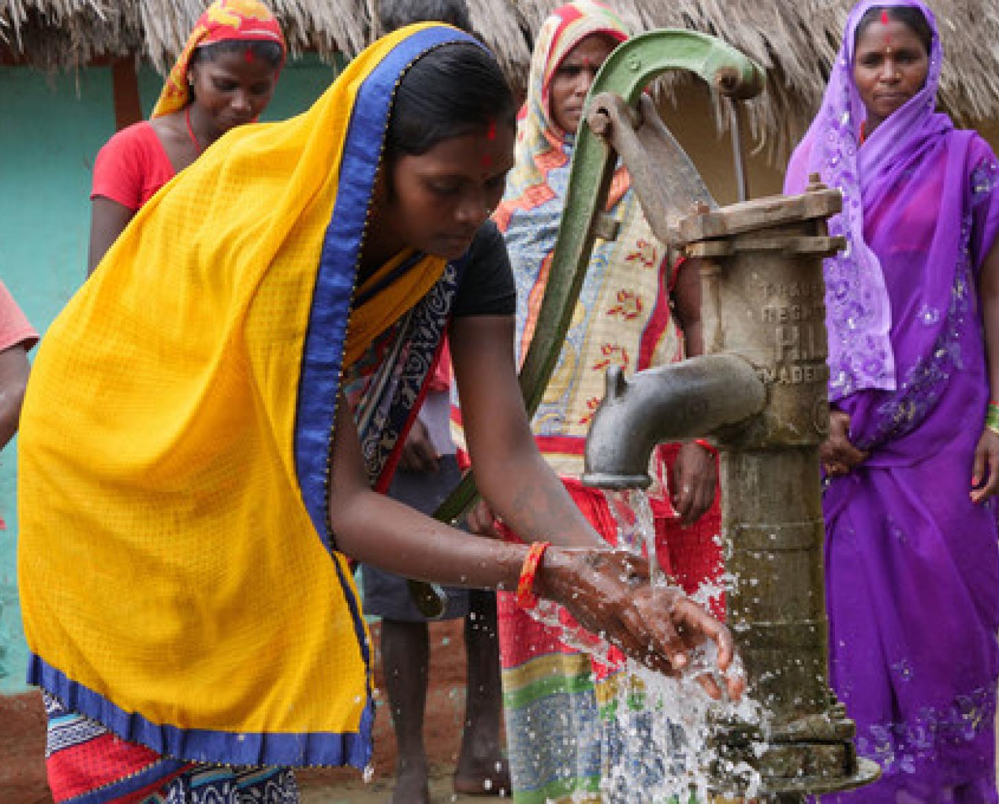 The embed image shows women and girls at a water pump washing their hands. 