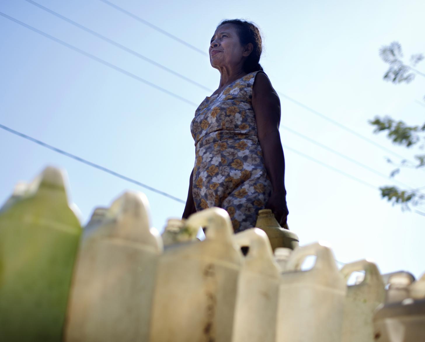 La mujer con gesto de determinación camina con un bidón de agua. En primer plano hay una fila de bidones de agua sobre el suelo.