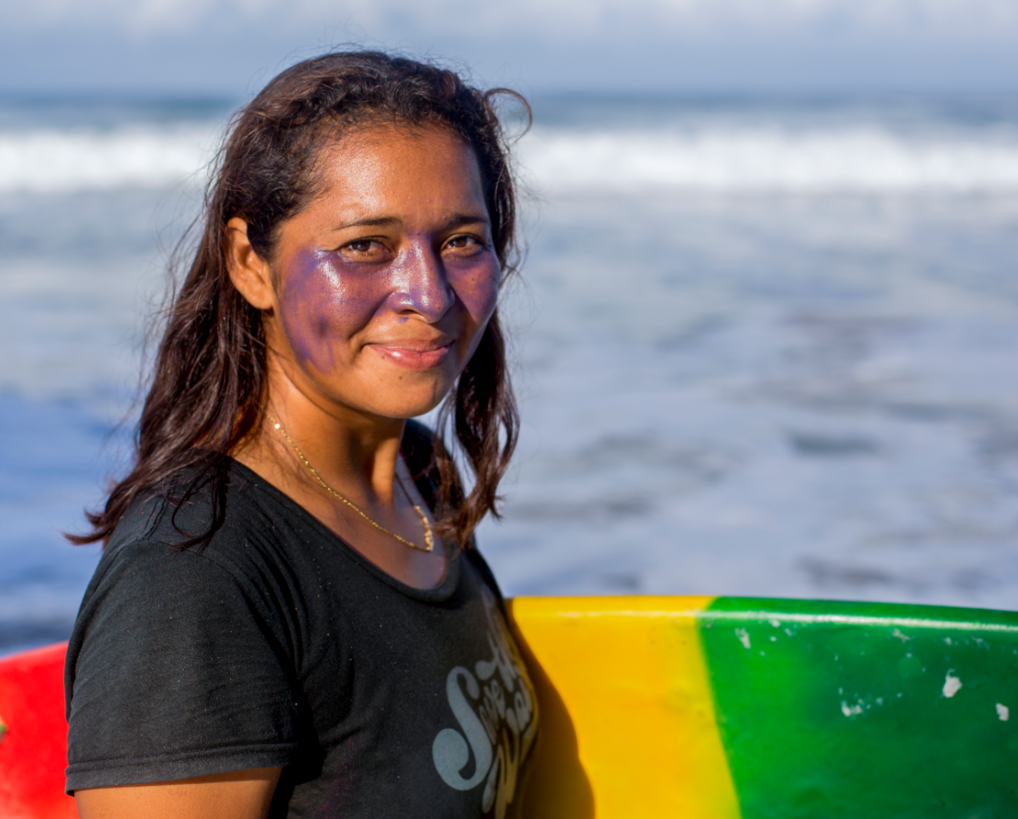 A female surfer smiles as she holds her surfboard by the shore of a beach.