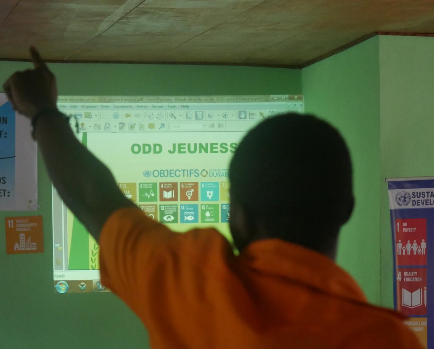 Un joven mira de frente a la cámara y levanta un dedo frente a una pantalla, en la cual los iconos de los ODS son proyectados.