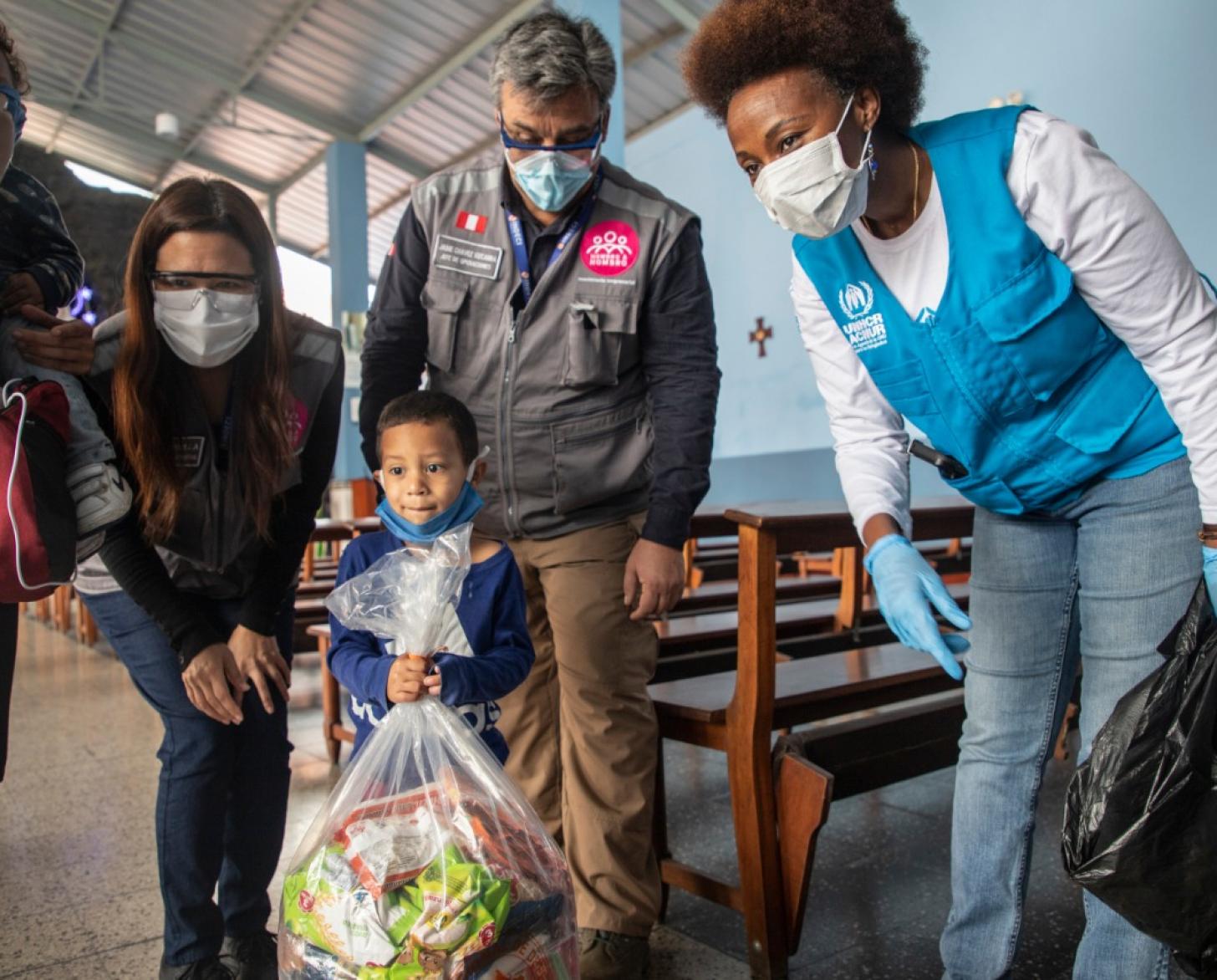 Personal de la ONU y de ONGs acompañan a un niño mientras sostiene una gran bolsa de alimentos y suministros donados.