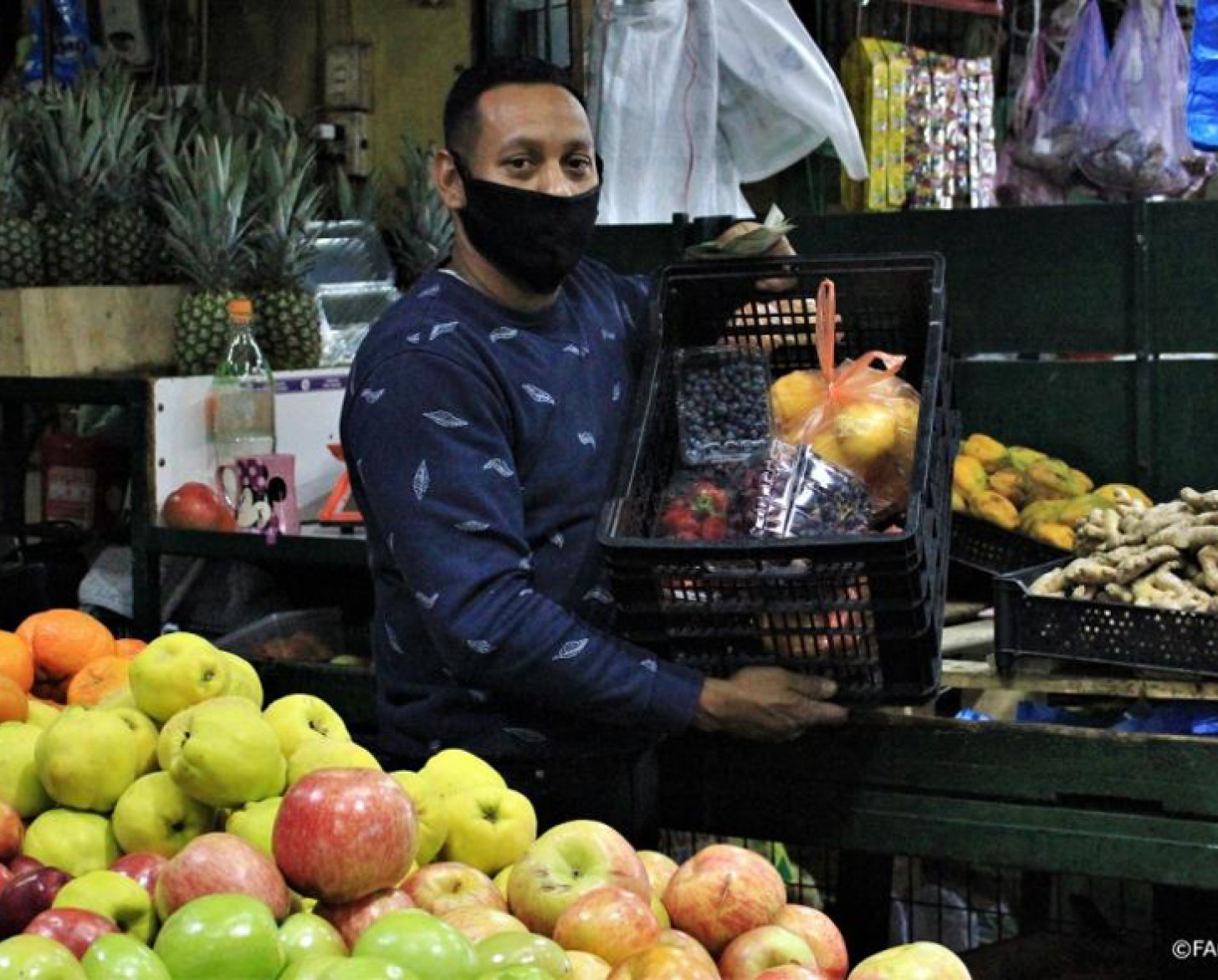 Un hombre con mascarilla negra sostiene una caja de frutas y verduras en un vendedor local de alimentos.