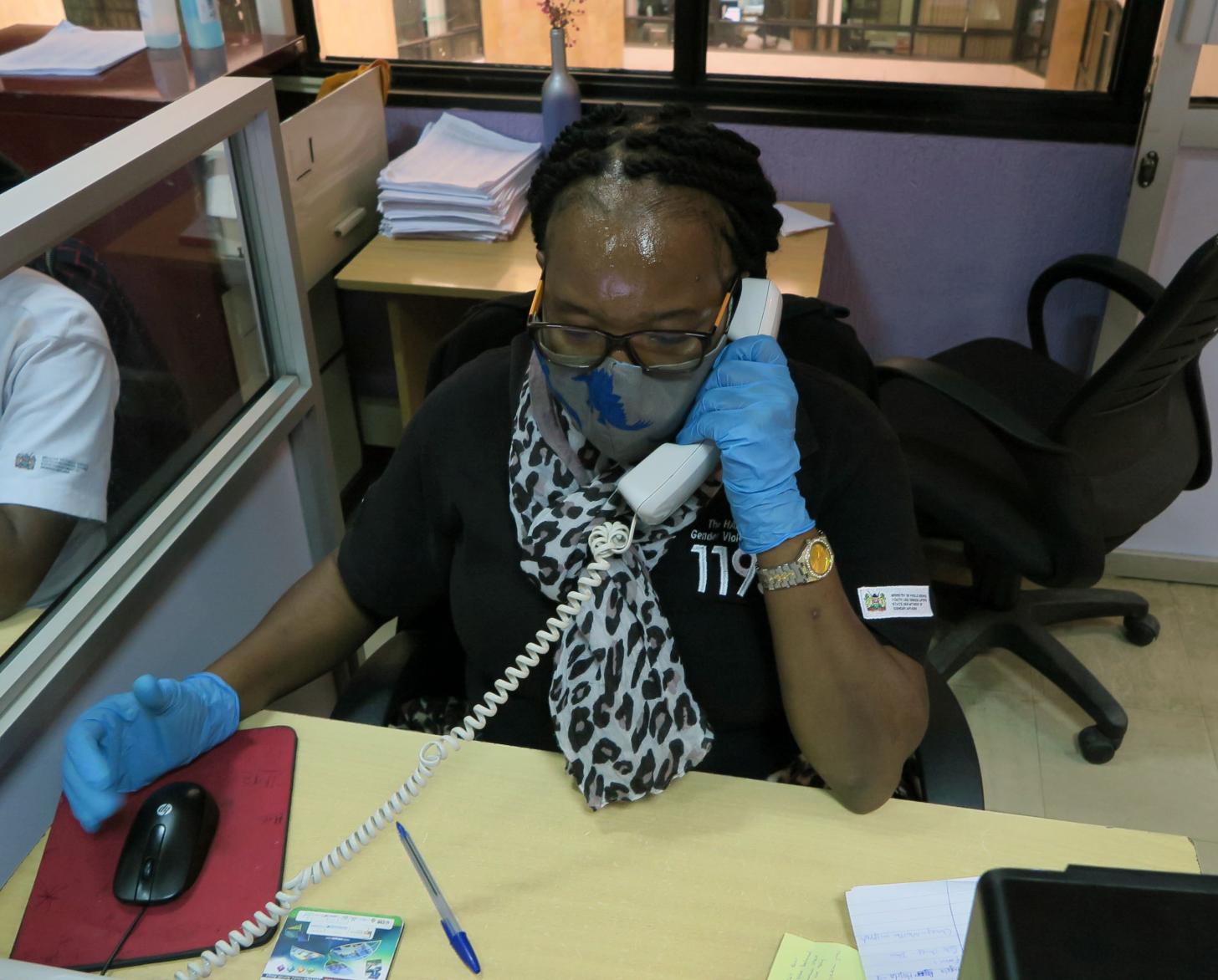 Une téléconseillère répond à un appel passé à la plateforme téléphonique du service d’aide aux victimes. Elle est assise à un bureau et porte un masque et des gants de protection.