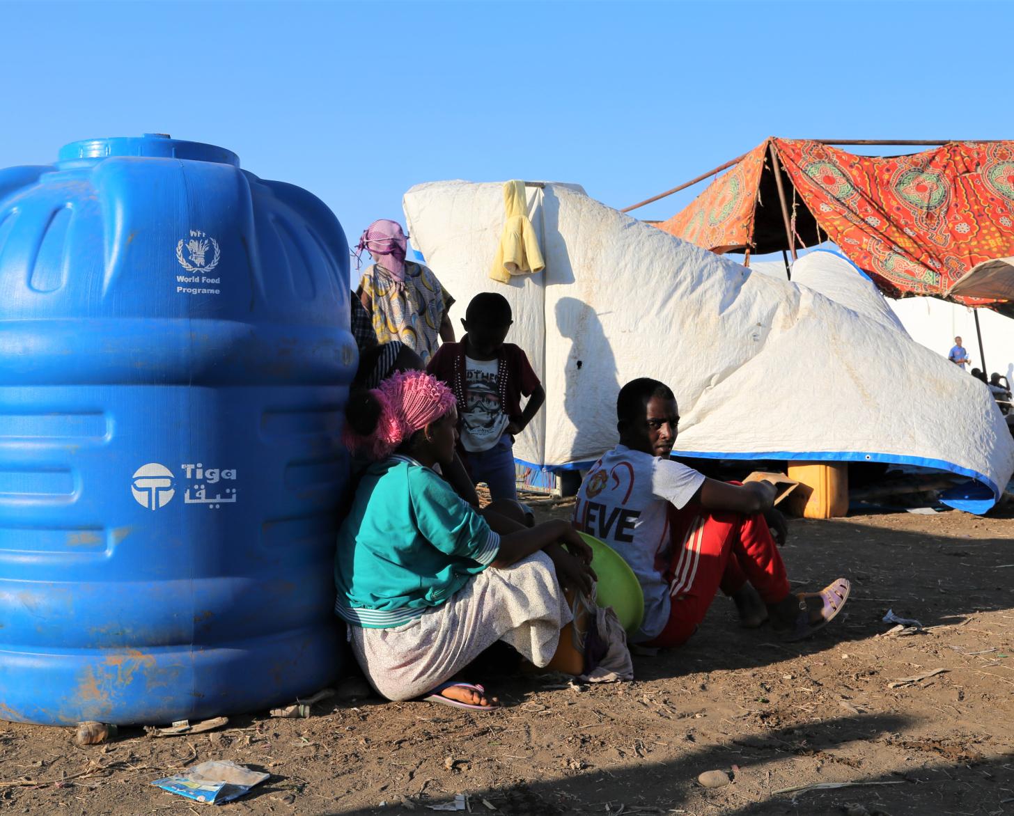 La gente se sienta frente a un gran contenedor azul de agua en el campamento.