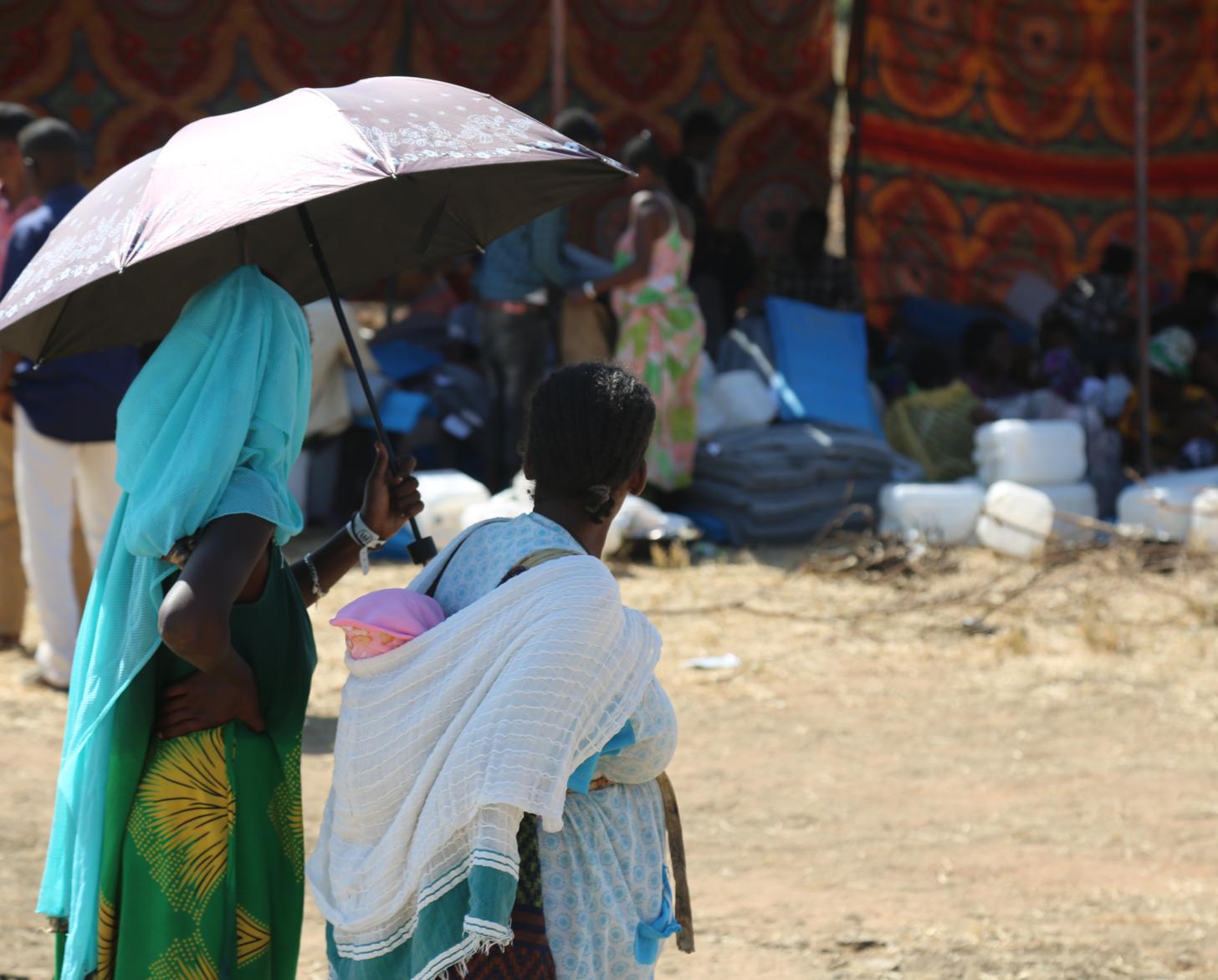 Dos mujeres (una con su hijo cargado) caminan bajo un paraguas en el campamento de refugiados.