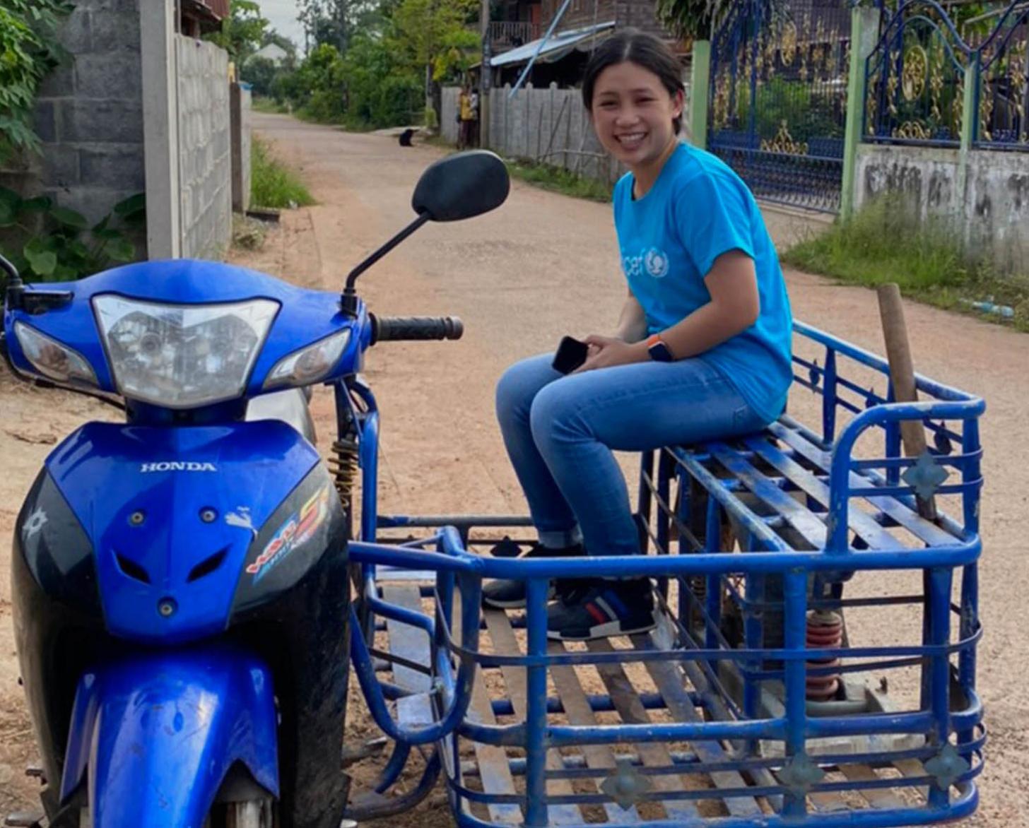 Une volontaire des Nations Unies vêtue d'un jean bleu et d'un t-shirt bleu portant le logo de l'UNICEF est assise dans le "side-cart" d'une moto bleue stationnée dans un chemin de terre et sourit à la caméra.
