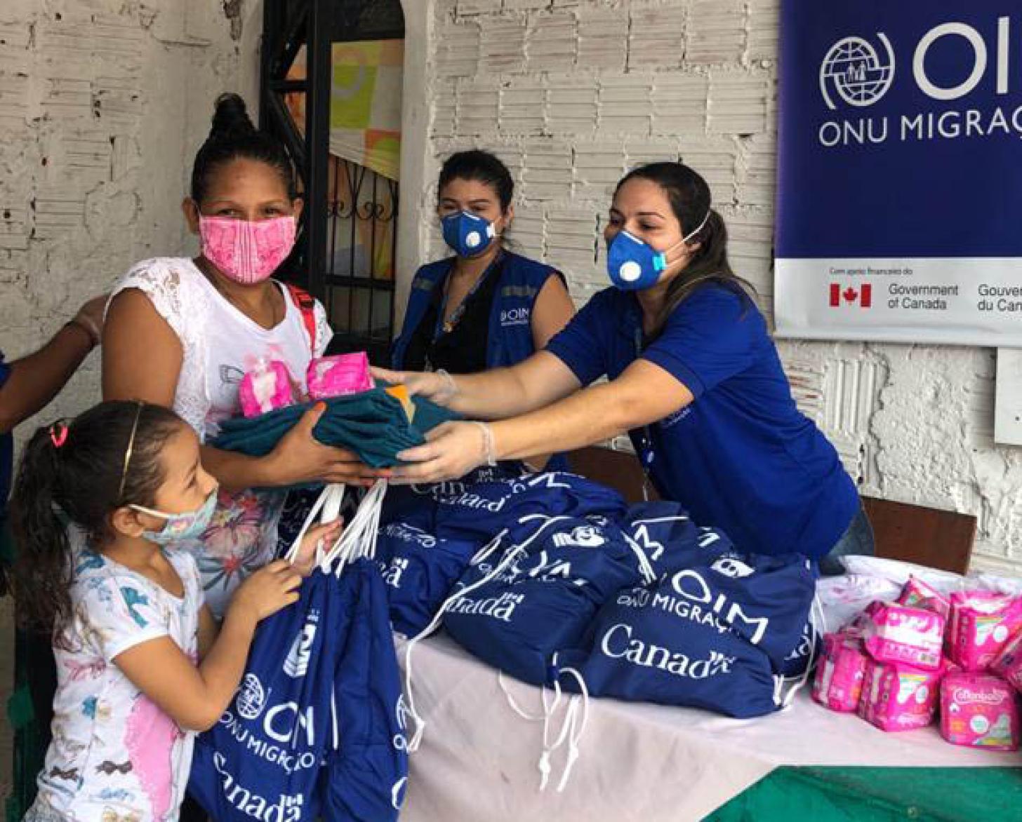 IOM staff a distribution station cheerfully hand over essential supplies to a mother and daughter.