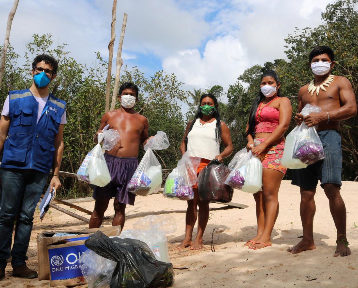 يقف موظف المنظمة الدولية للهجرة مرتديًا كمامة قرب عائلة من السكان الأصليين بعد تلقيها المساعدات.