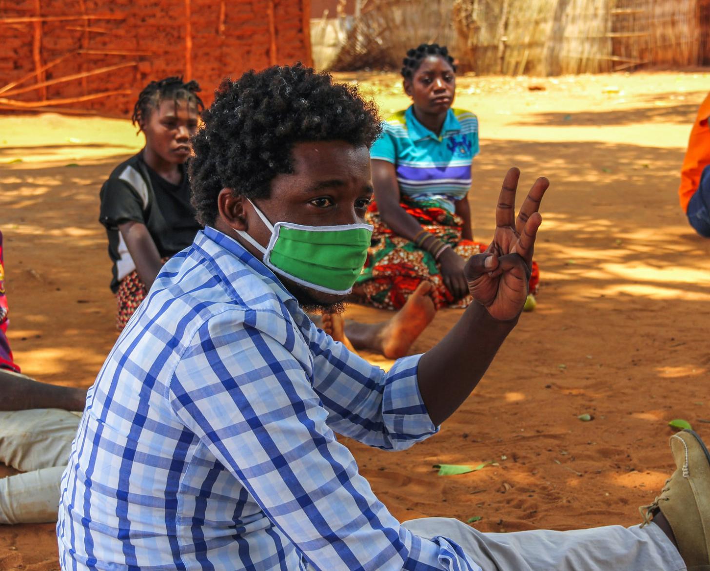 رجل في وسط الصورة يرتدي كمامة ويرفع ثلاثة أصابع بينما يجلس آخرون في نصف دائرة في الخلفية.