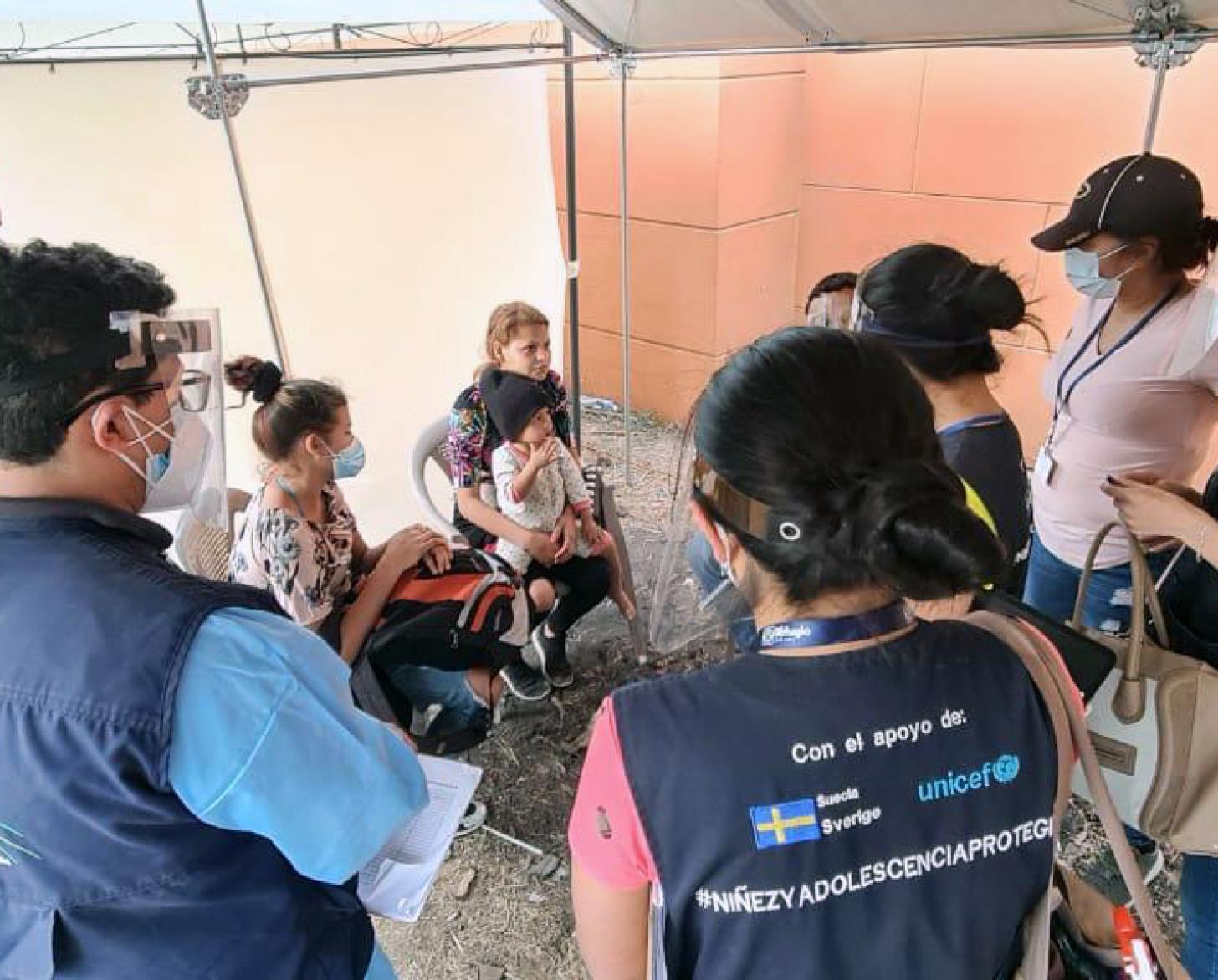 يقف موظفو الأمم المتحدة الذين يرتدون كمامات ودروع واقية للوجه حول أسرة جالسة، وكل ذلك تحت خيمة.