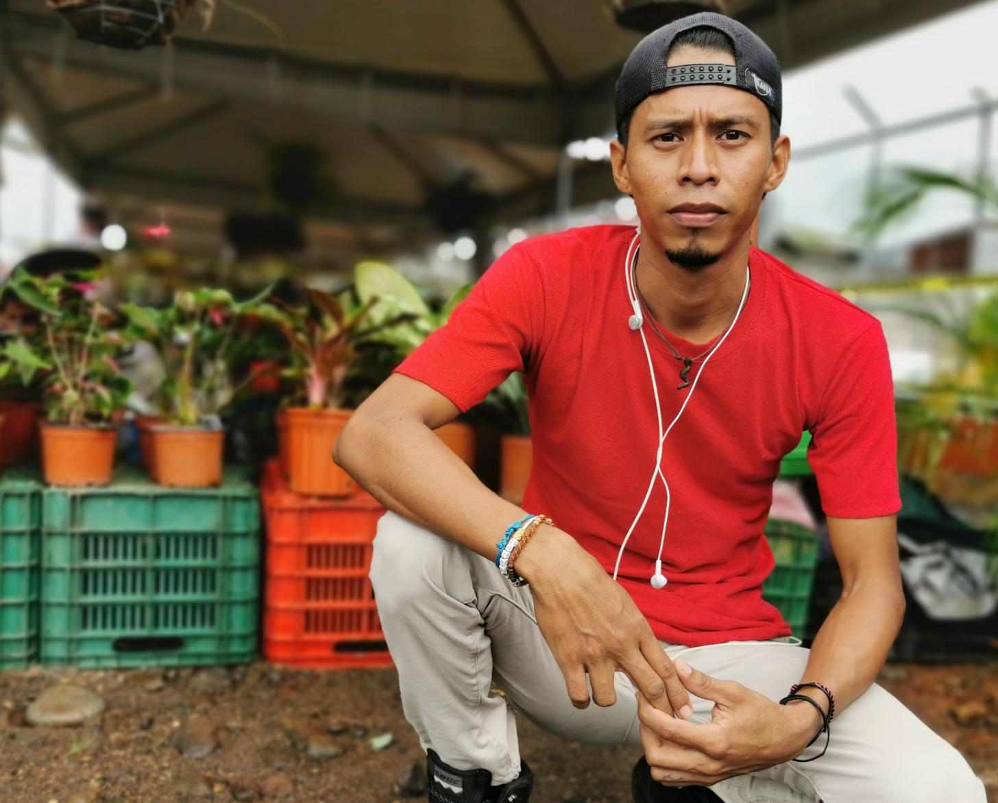 Juan Carlos kneels on the ground under a tent and in front of a row of plants. 