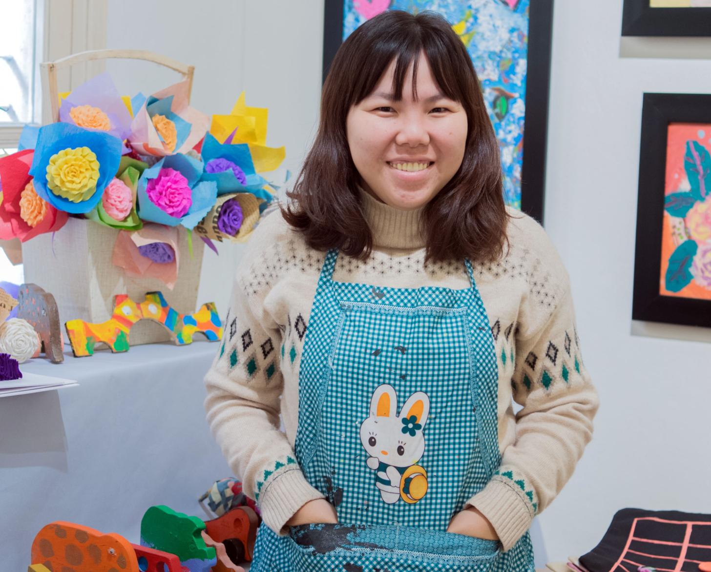 Tu Thanh Thuy smiles brightly at the camera as she stands with her hands in the pockets of her apron in front of colourful paper flowers and paintings.
