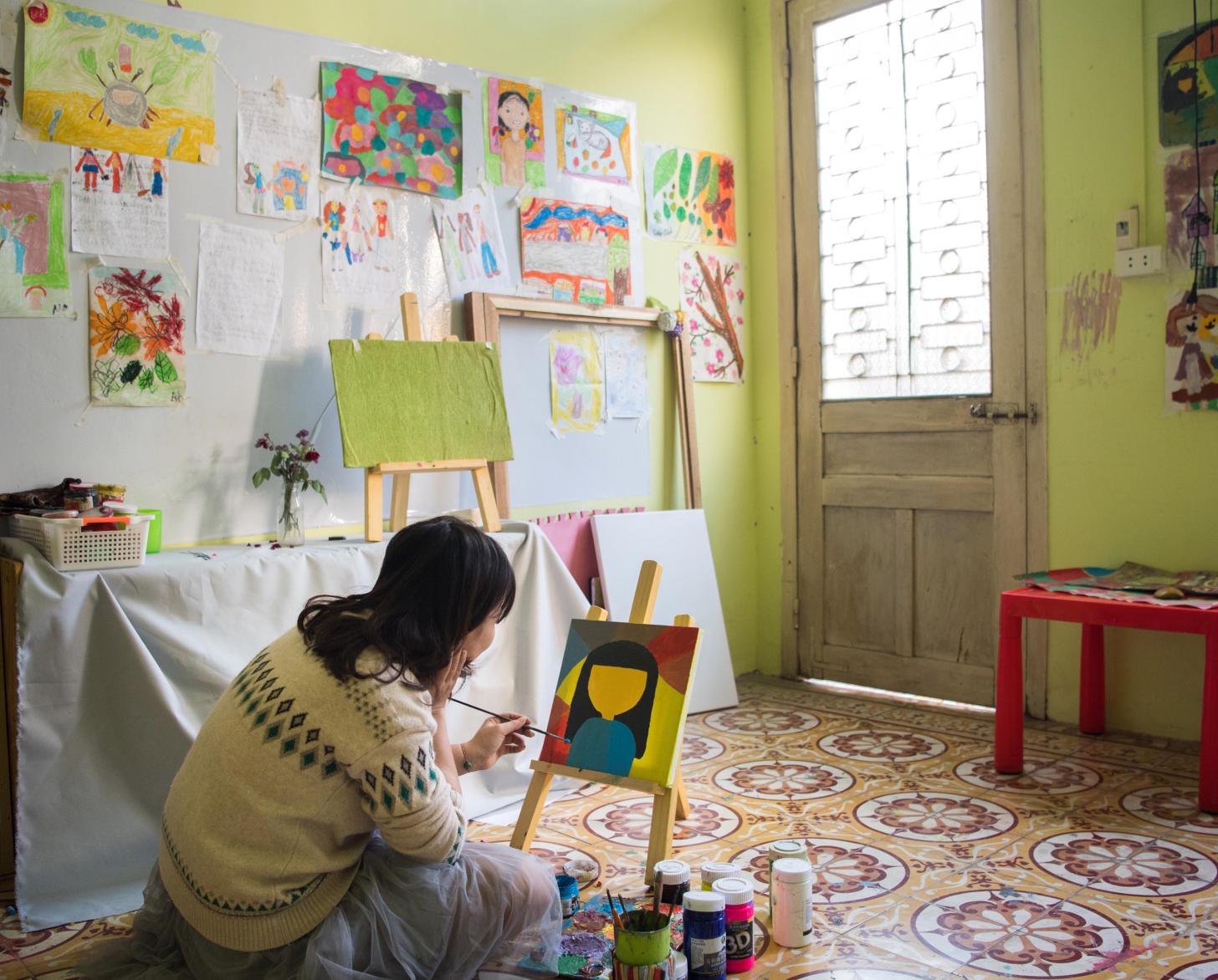 Tu Thanh Thuy sits on the floor of an art space and paints a picture placed on a small canvas.