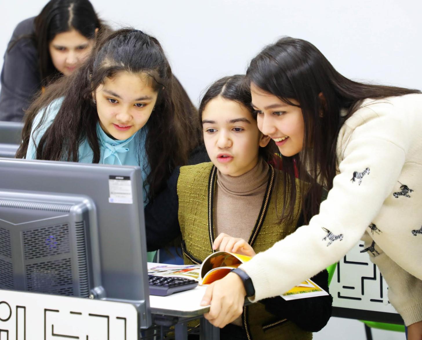 Группа улыбающихся девушек вместе работают за компьютером. Еще одна девушка на заднем плане работает за компьютером. 