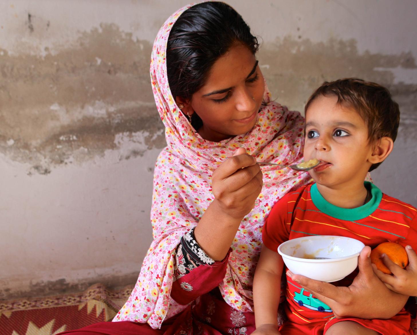 A young woman holds a child in her lap while she feeds him from a bowl.