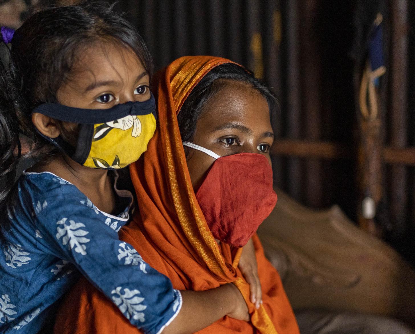 一个戴着面罩的小女孩从后面抱着她的母亲，她的母亲坐在那里。