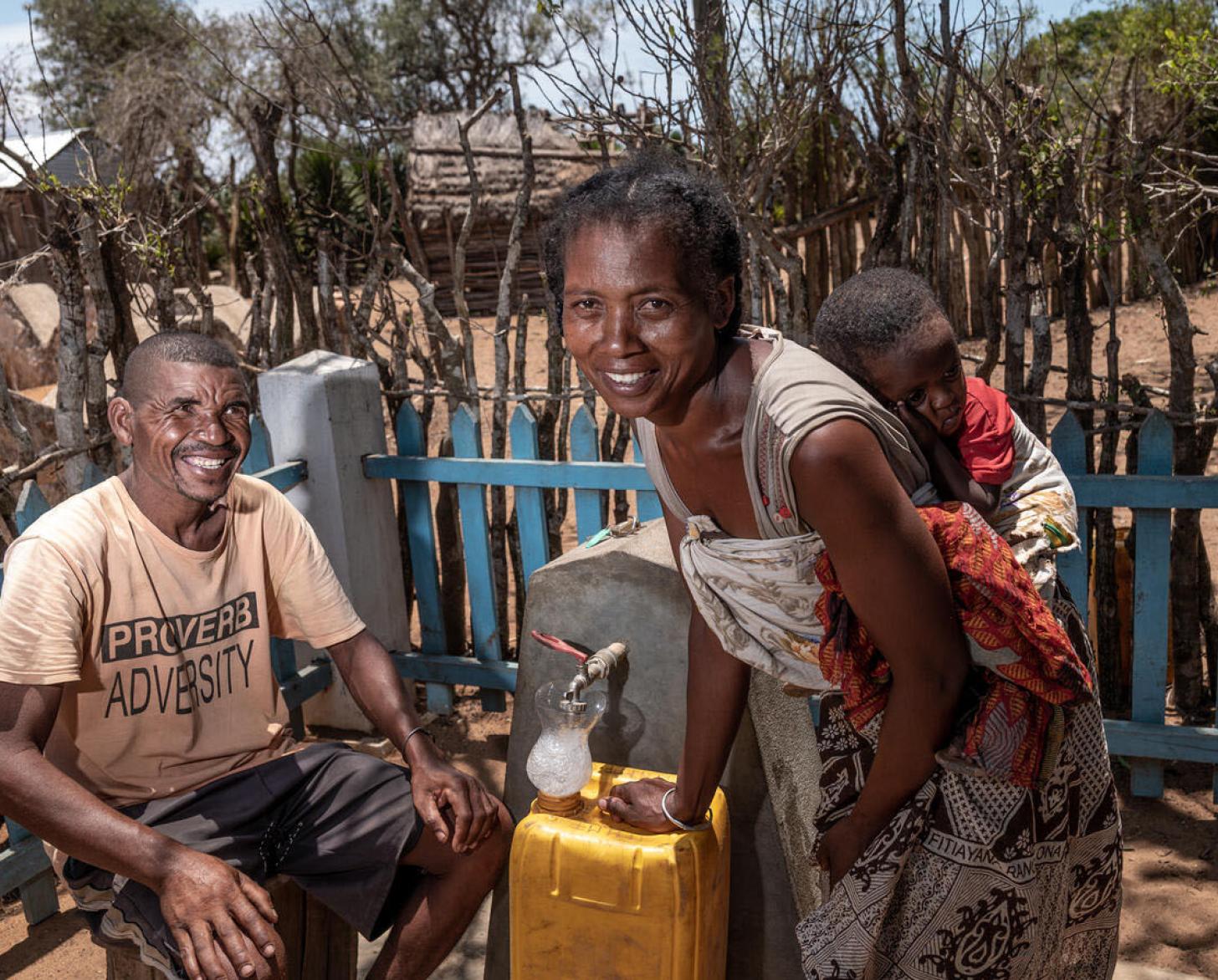 A woman carrying her sleeping baby on her back and a man smile cheerfully while next to their water fountain.