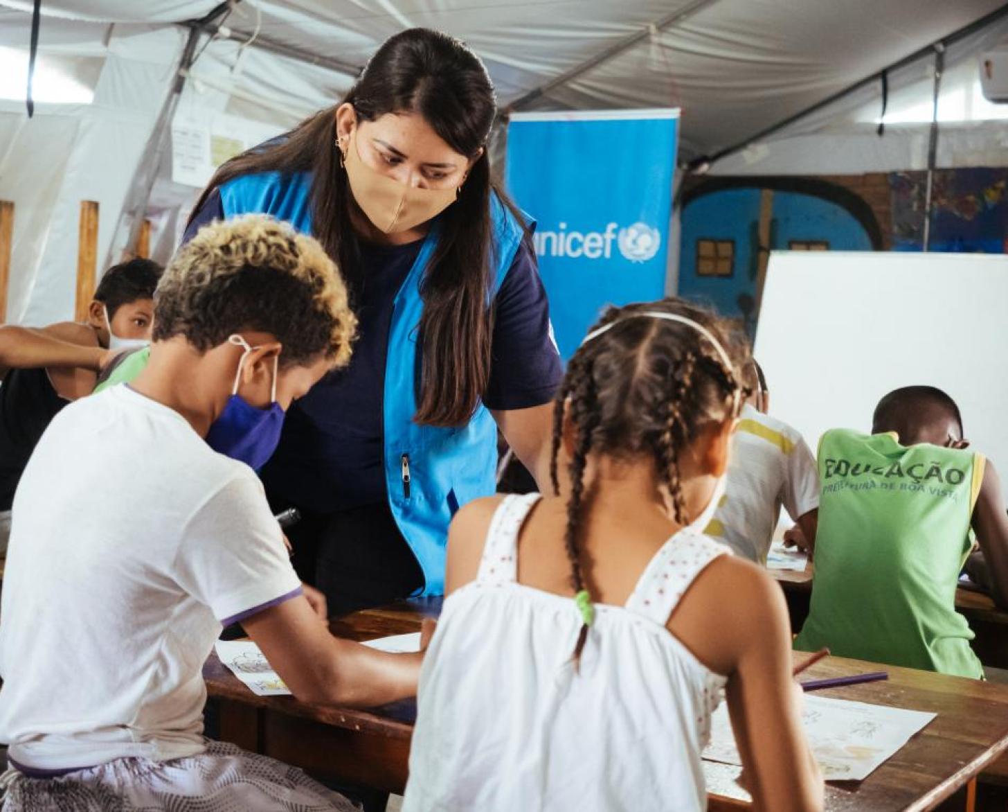 A teacher with a face mask and blue vest talks to a boy and a girl in a classroom. 