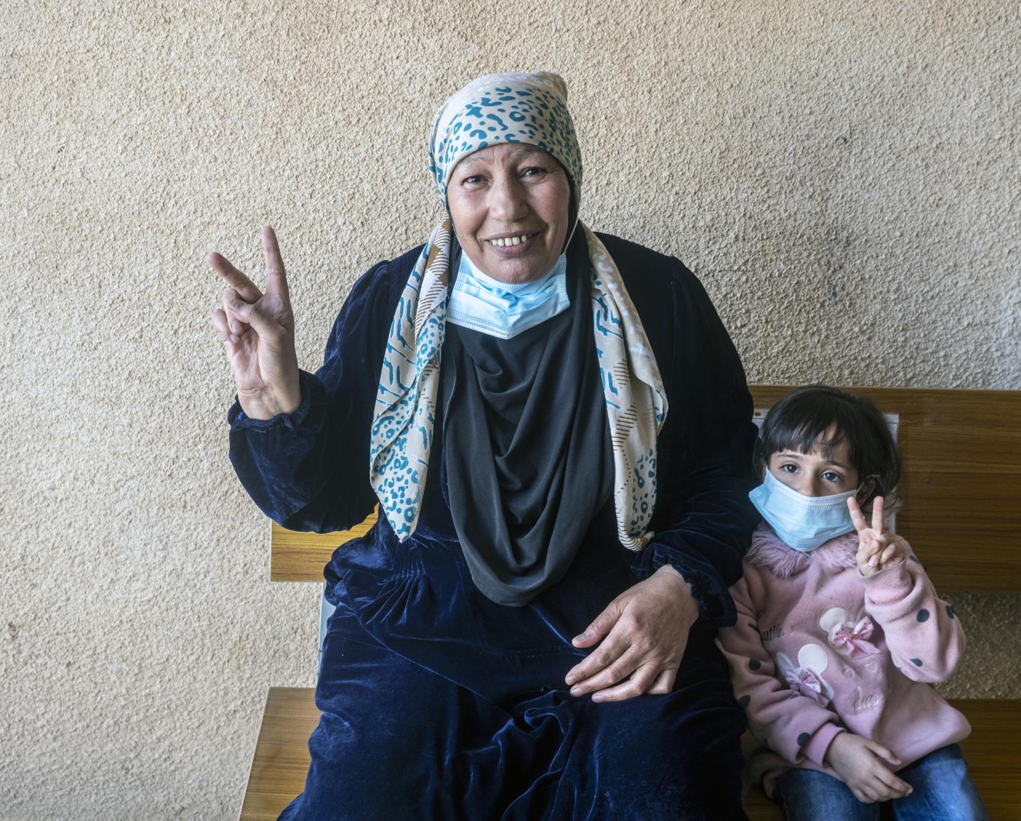 A woman smiles cheerfully at the camera with her young daughter. Both flash the 'V' for vaccine sign with their fingers.
