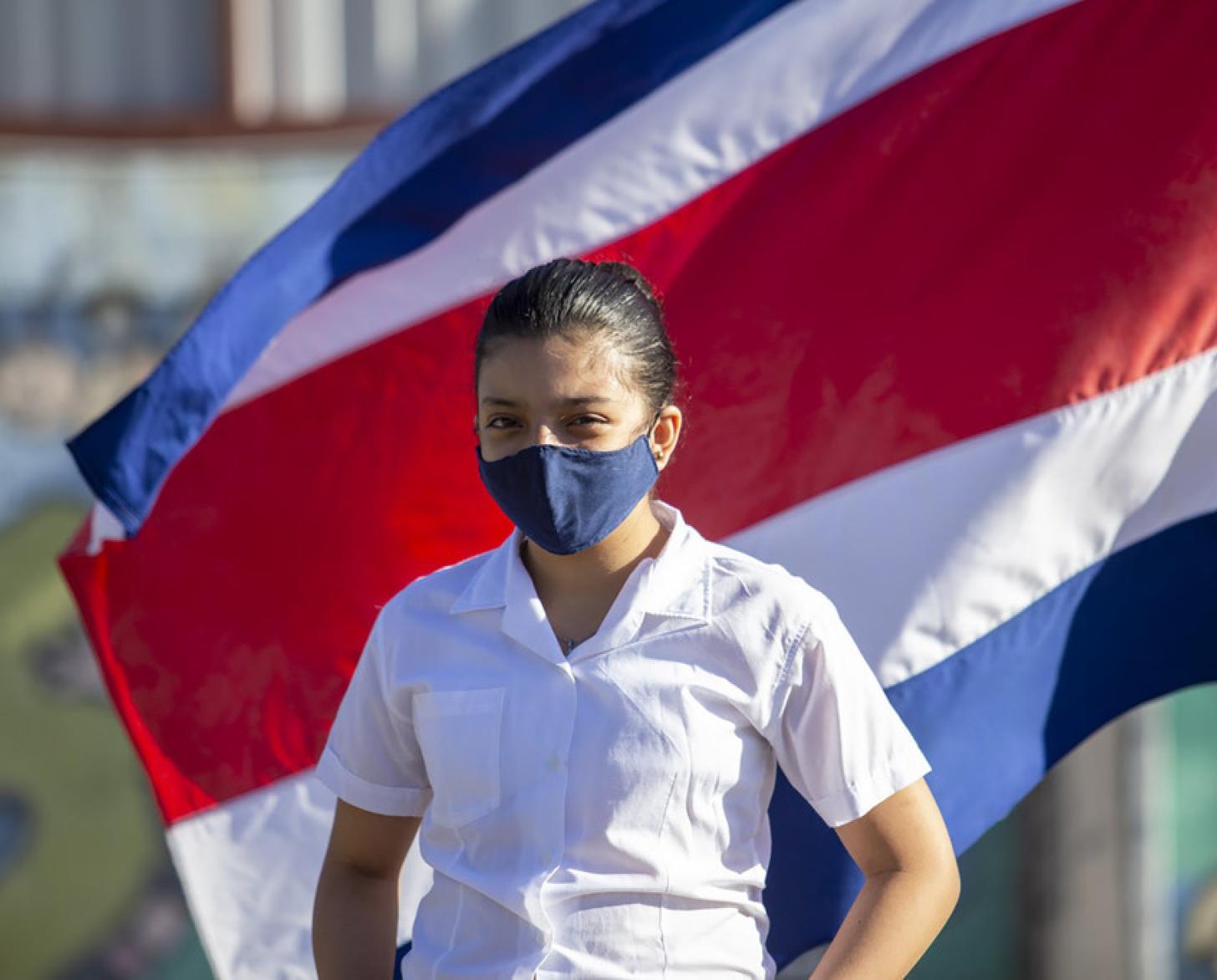 فتاة ترتدي قناعًا أزرق تقف أمام علم كوستاريكا.