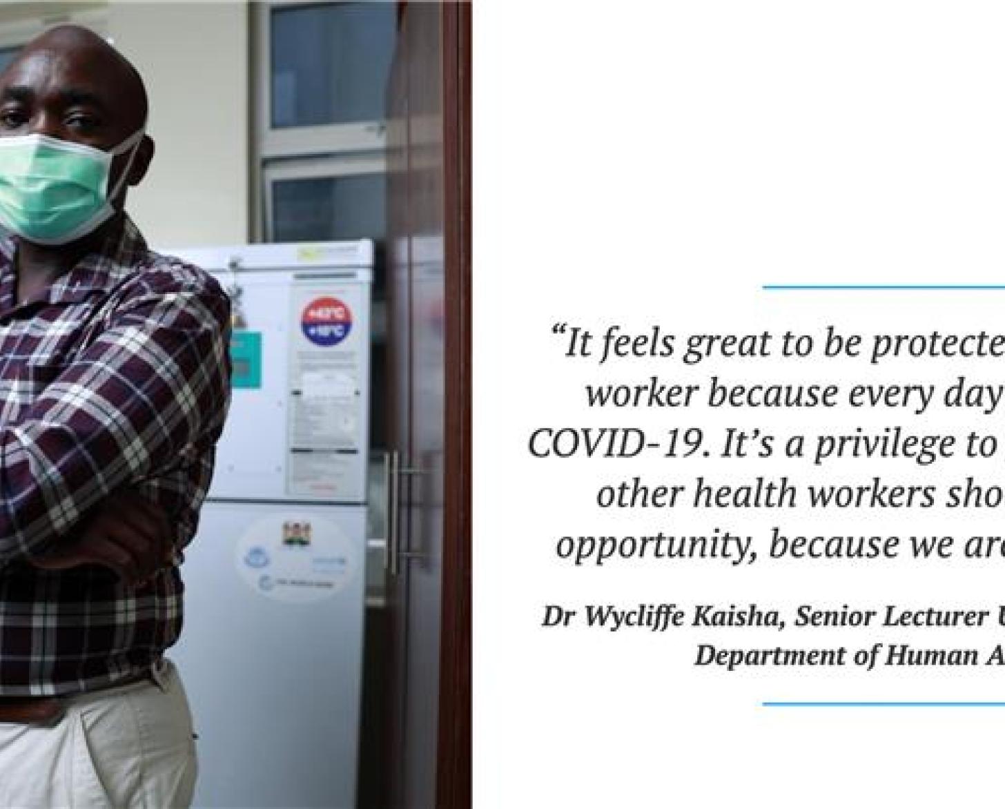 A man in a plaid shirt and a green face mask has his arms crossed while looking at the camera. 
