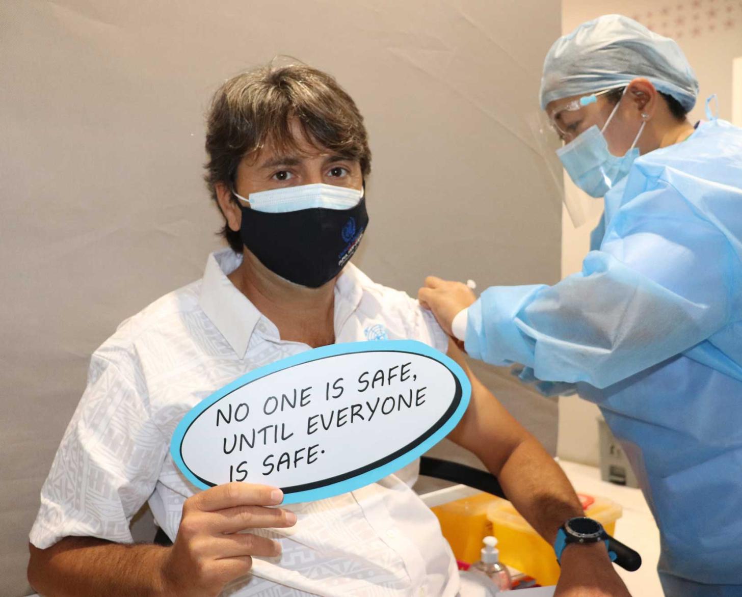 The Resident Coordinator of Papua New Guinea is shown getting his COVID-19 vaccine as he proudly displays a sign noting the safety of the vaccine.