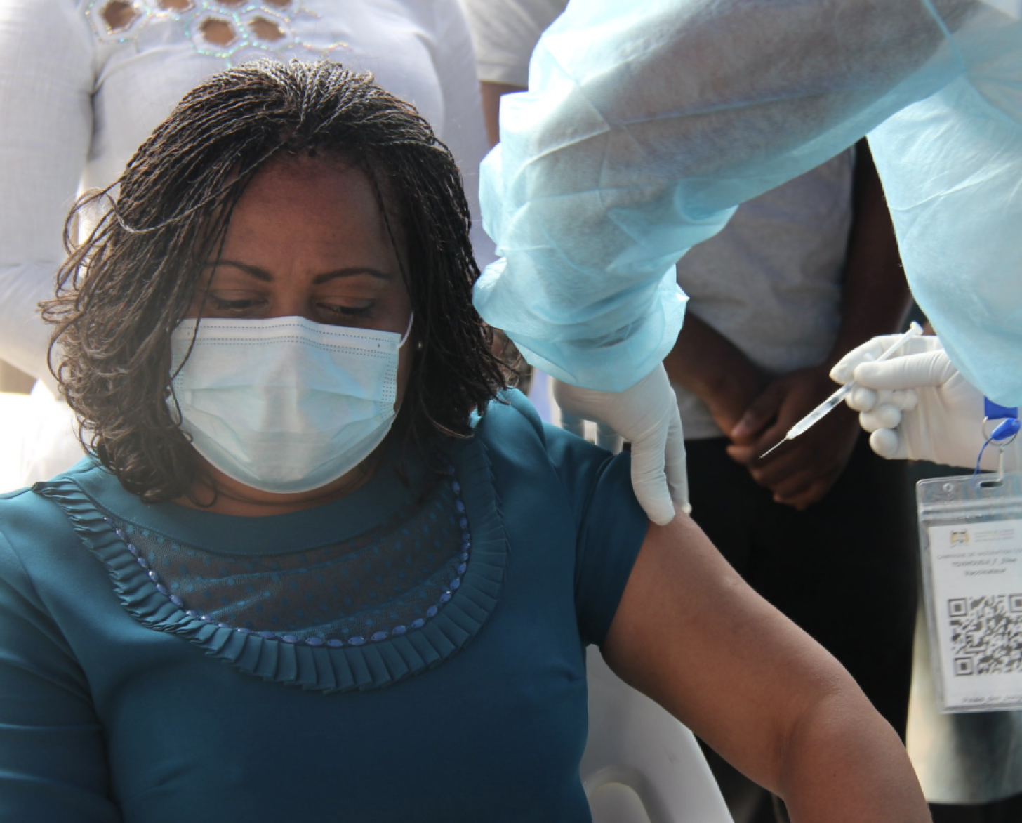 امرأة ترتدي قميصًا أزرق مخضر وتنظر إلى الأسفل بينما تتلقى اللقاح من قبل شخص يرتدي معدات واقية.