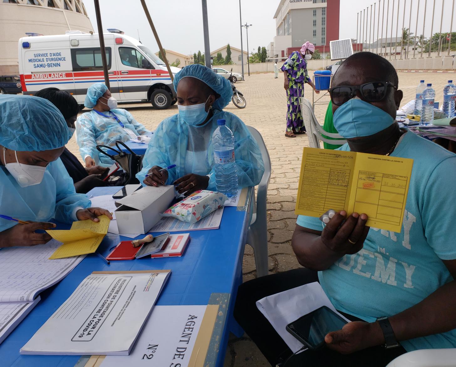 رجل يرتدي نظارة شمسية وقميصًا أزرق وكمامة يظهر استمارة التطعيم أمام الكاميرا بينما يجلس اثنان من العاملين في مجال الرعاية الصحية بالقرب منه.