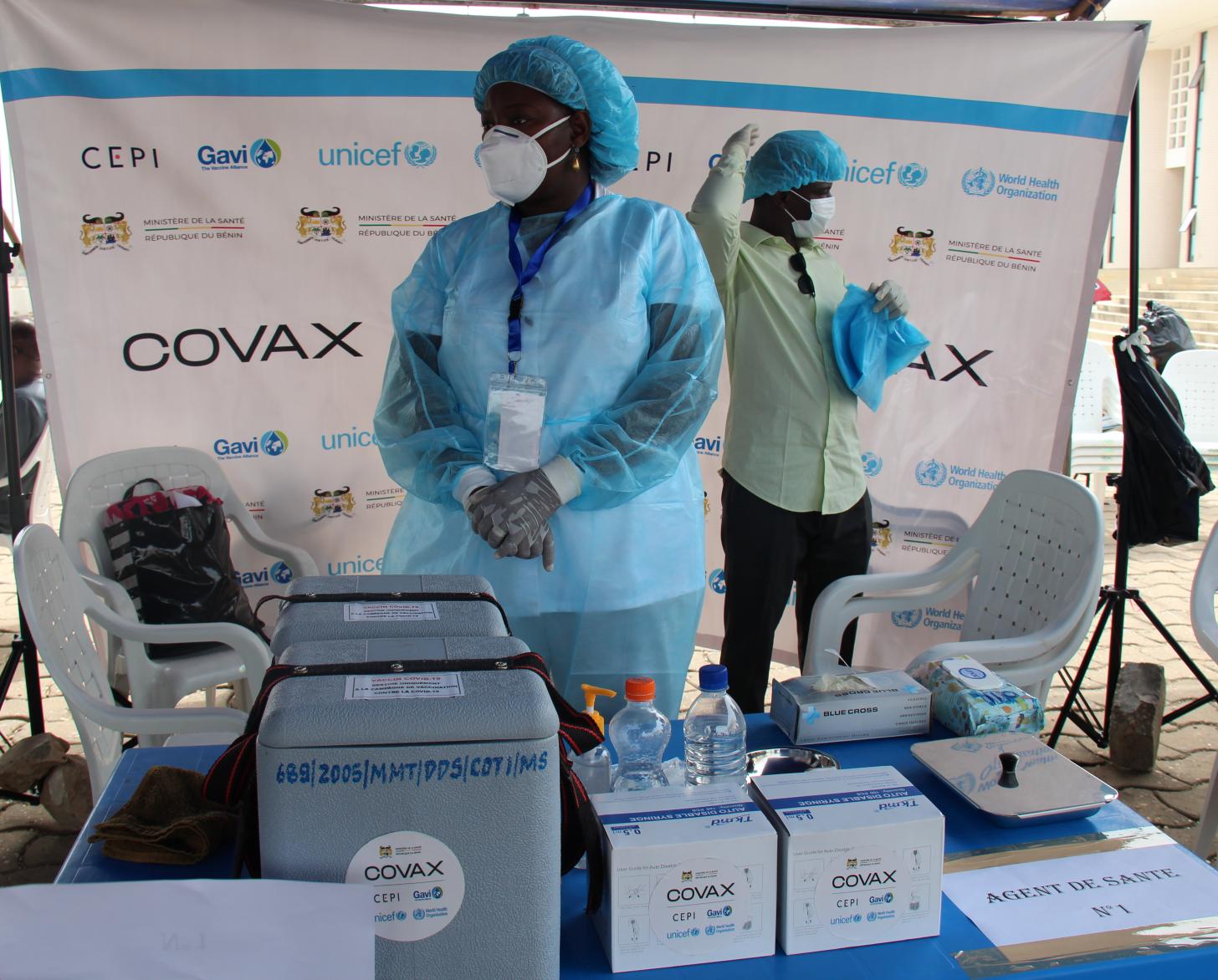 شخصان يرتديان معدات طبية وقائية يقفان بالقرب من إمدادات التطعيم.