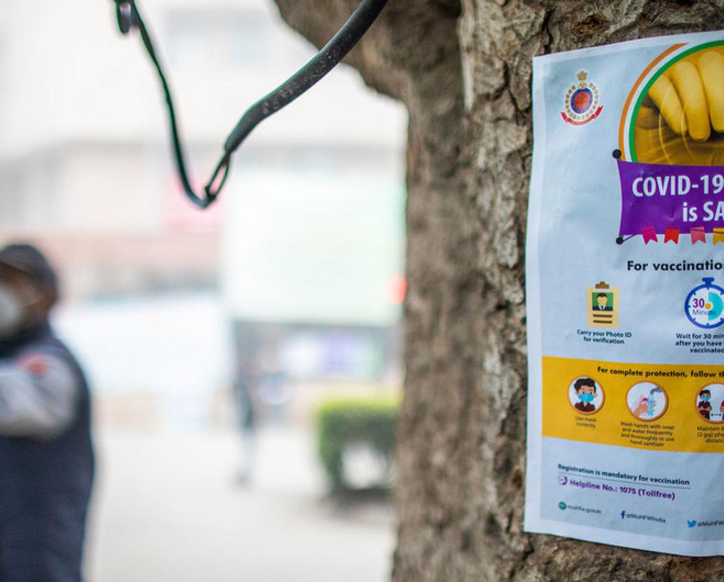 A sign, with the image of a vaccine and information, is posted on a tree and a person in a mask is seen in the background pointing. 