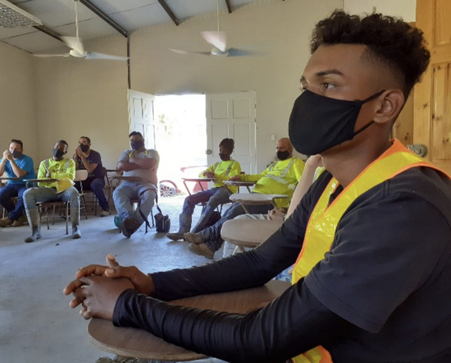 يجلس رجال عدة على مكاتب بالقرب من رجل يرتدي قميصًا أسود وسترة بناء ينظر إلى مكبر صوت.