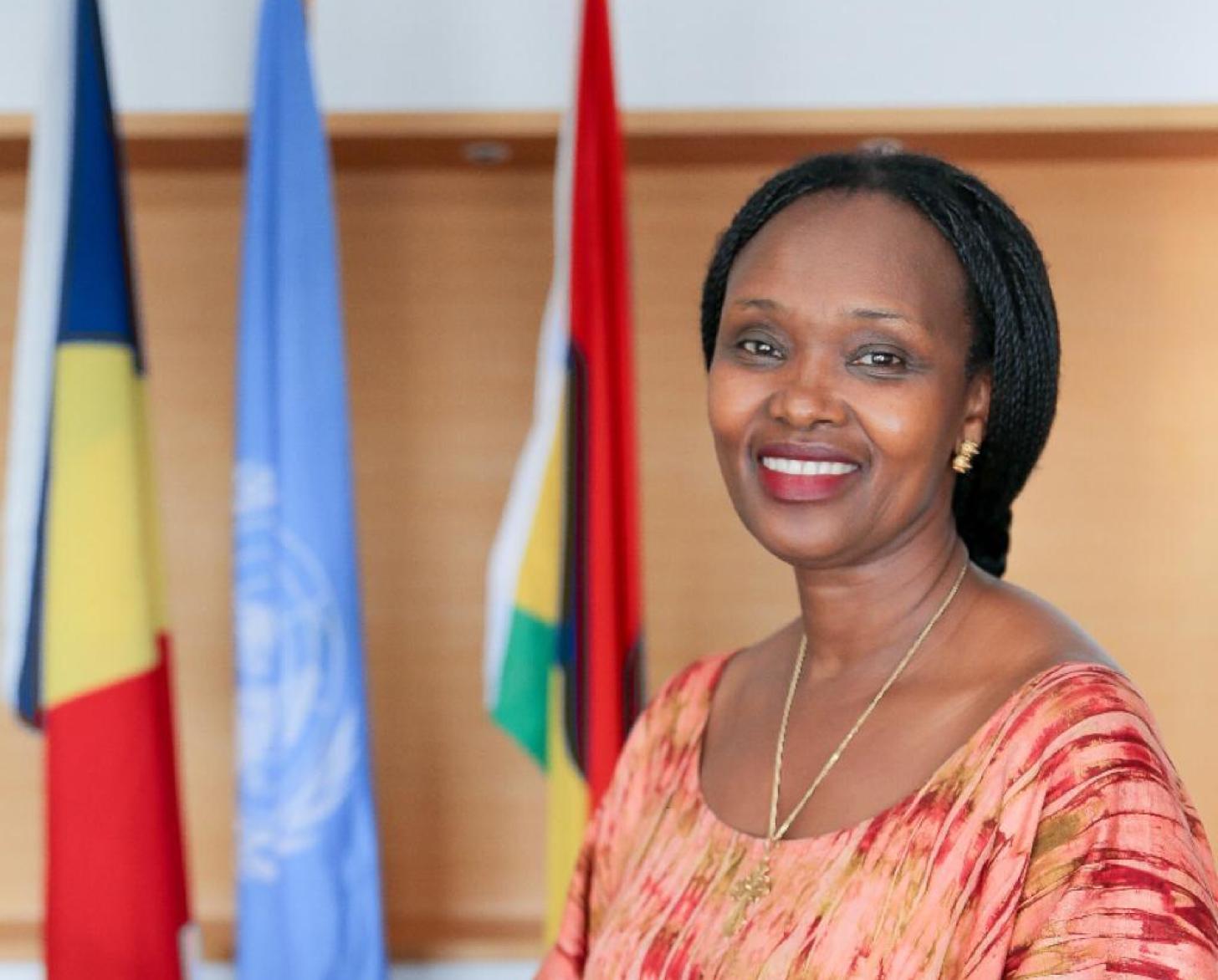 Una mujer con una camisa naranja y roja sonríe a la cámara frente a tres banderas.