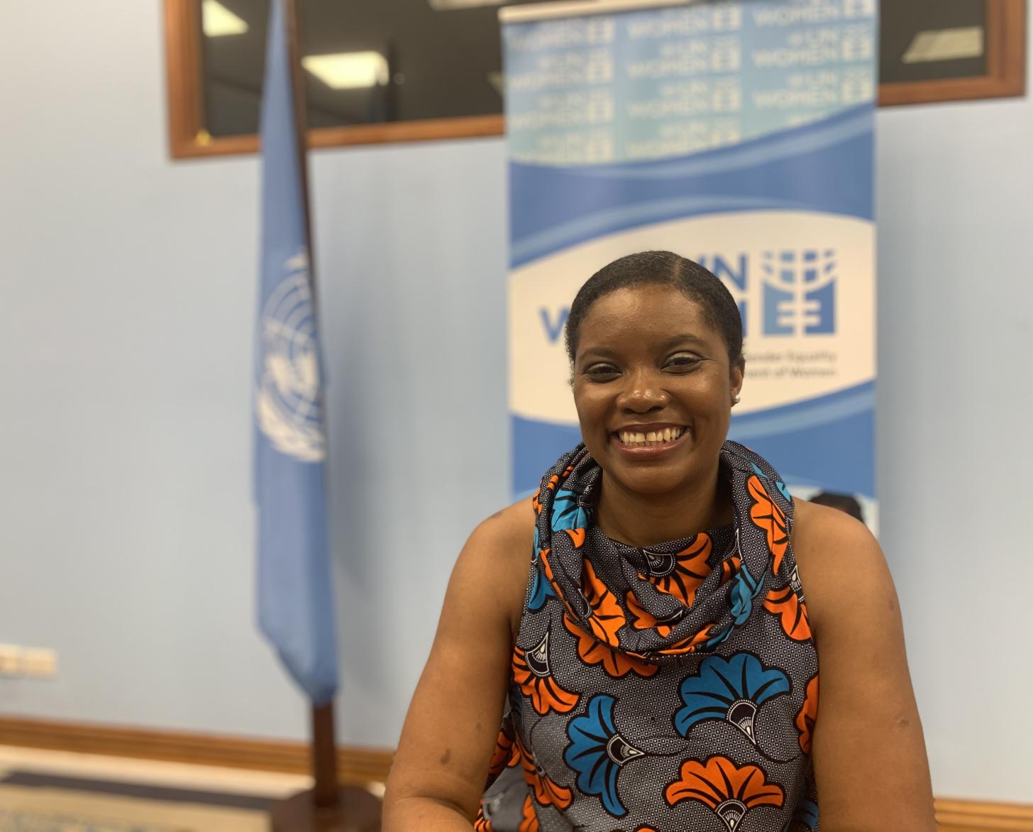 Una mujer con una camisa gris con flores sonríe a la cámara frente a una pancarta de ONU Mujeres.