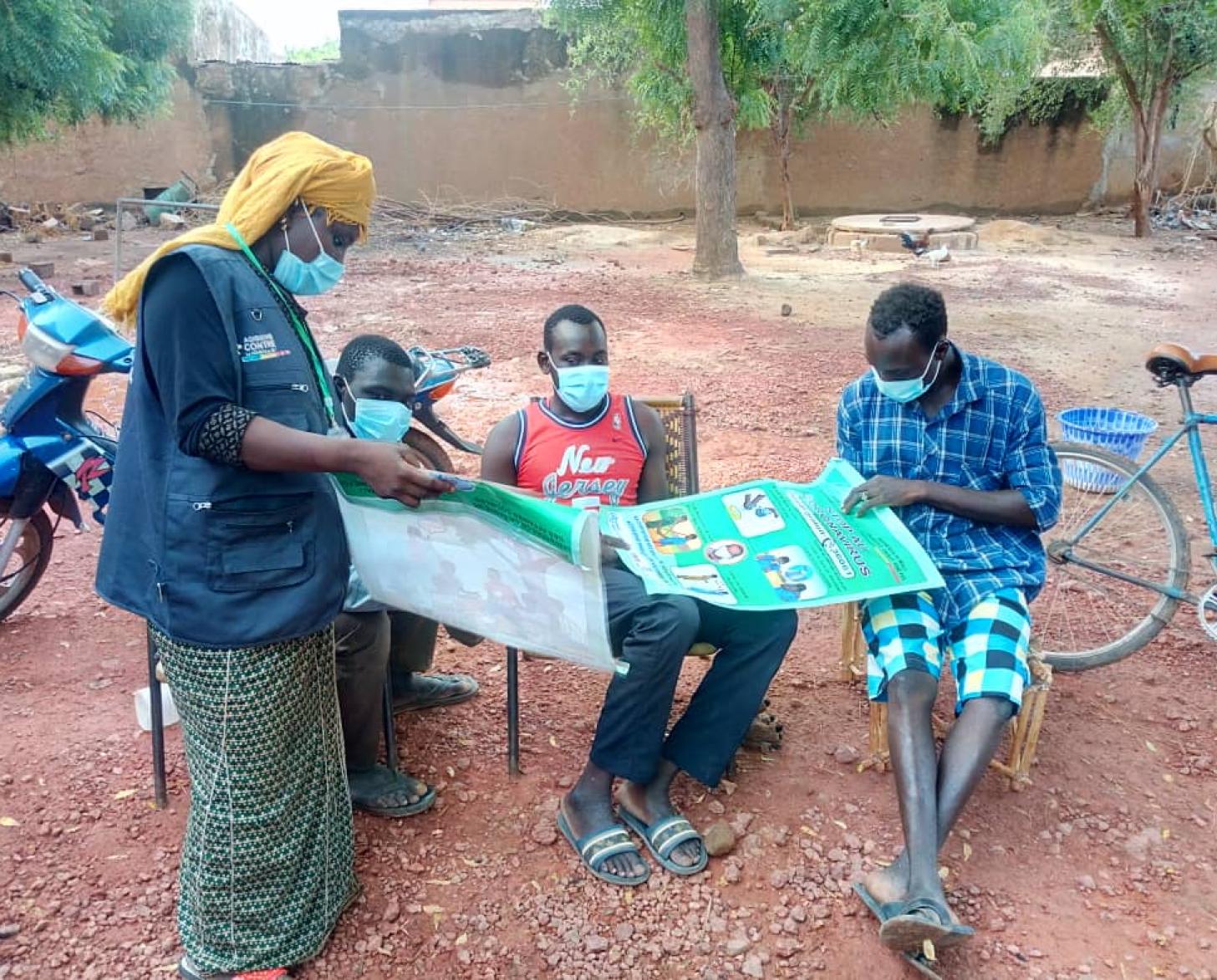 One person standing over three people showing a large poster. Another man holds the poster in his hand and reads it.