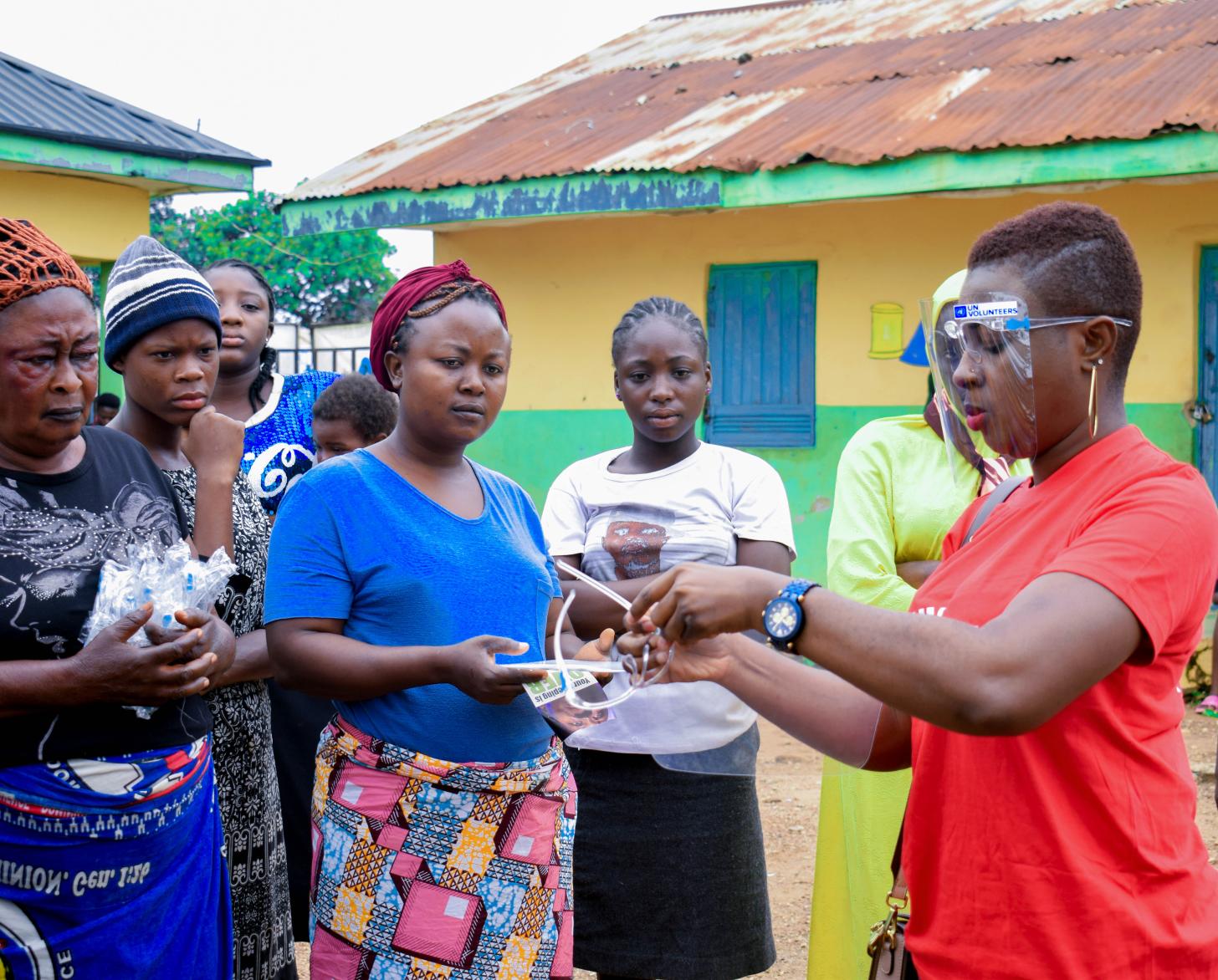 A woman in a red shirt and face shield shows a group of women and girls a pair of goggles.