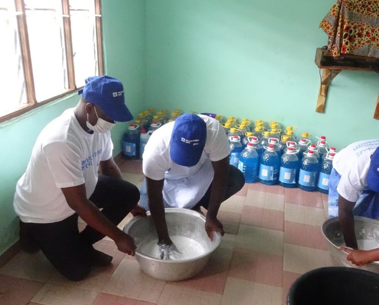 Four people kneel on the ground with two metal pails. Two individuals have their hands in the pails and are mixing up a white substance.