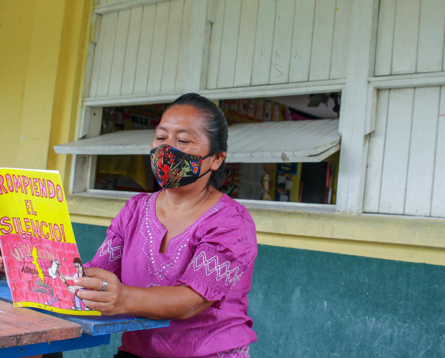A woman in a colorful face mask and purple shirt reads a book while sitting at a table.